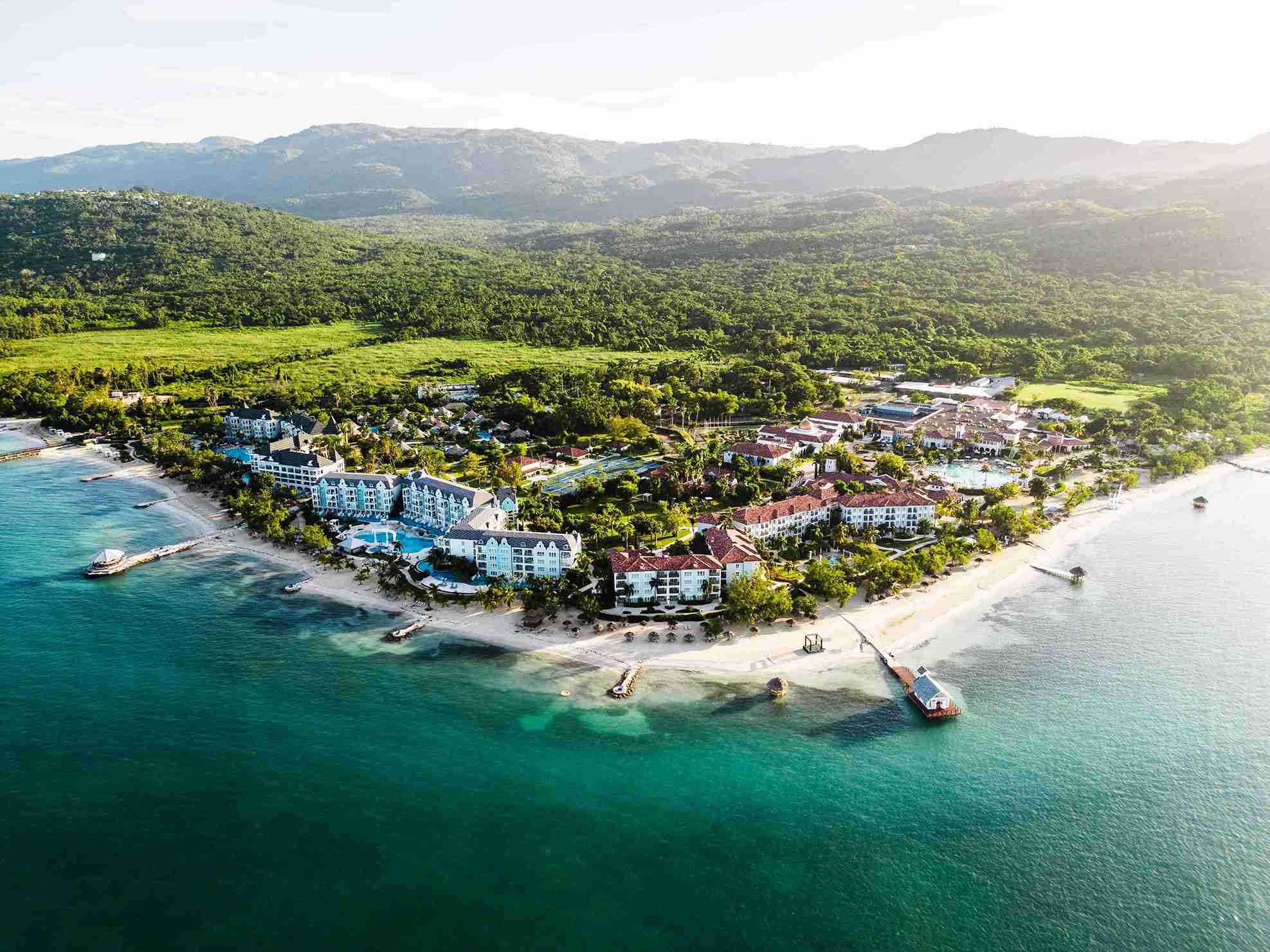 Aerial view of a coastal resort surrounded by greenery, featuring white buildings with red roofs, pools, and sandy beach along a turquoise sea with boats anchored nearby.