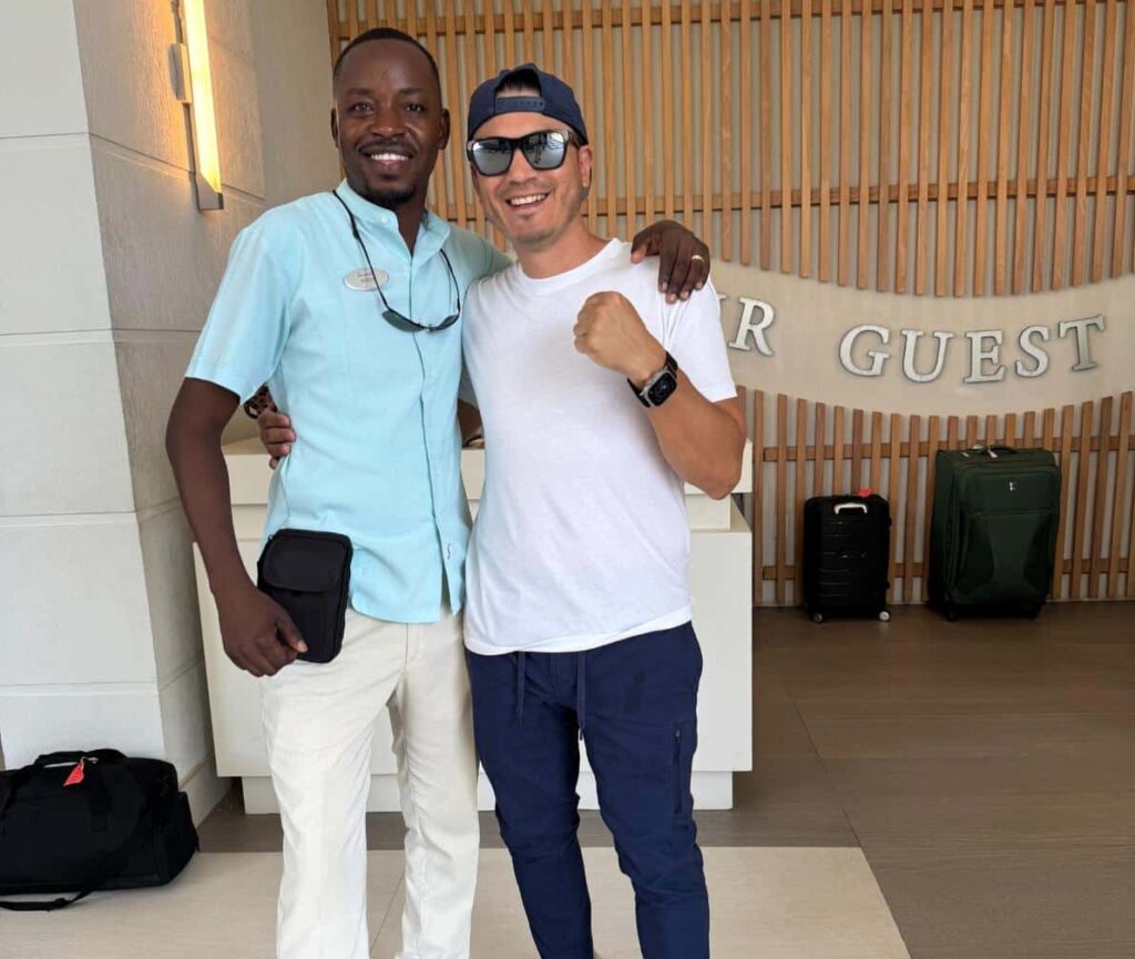 Two men stand together in a welcoming hotel lobby. One man wears a light blue short-sleeve shirt and beige pants with brown shoes and a name tag, smiling confidently. The other man is dressed in a white t-shirt, blue pants, and sneakers, giving a thumbs-up with a joyful expression. In the background, the hotel check-in area features a wooden slatted wall and luggage.