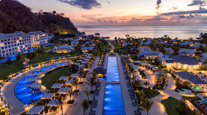 Aerial view of a luxury resort at sunset, featuring multiple swimming pools and cabanas surrounded by palm trees, with the ocean and distant islands in the background.