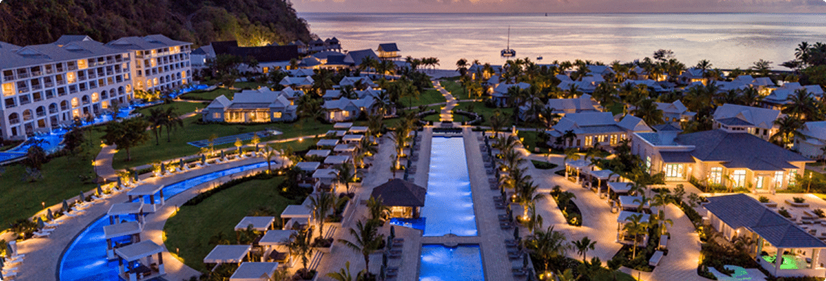 Aerial view of a luxury resort complex at dusk, featuring illuminated pathways, swimming pools, and lush gardens, with the ocean visible in the background.