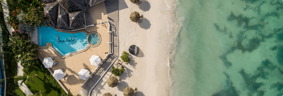Aerial view of a sandy beach with a pool area featuring umbrellas and lush greenery, adjacent to clear turquoise waters.
