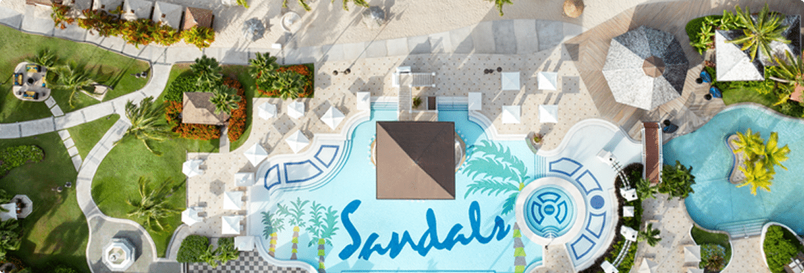 Aerial view of a resort pool area with the word 'Sandals' painted in blue on the bottom of the pool, surrounded by lush greenery, beach loungers, white umbrellas, and cabanas.