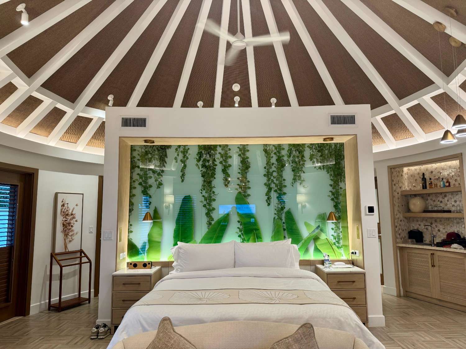 A stylish bedroom with a circular ceiling featuring wooden beams. In the center, a large bed is dressed in white linens, flanked by nightstands. Behind the bed, a focal wall with a green glass panel adorned with hanging plants and decorative lighting creates a modern, nature-inspired ambiance. To the left, there's a doorway with a wooden door leading to another room, and to the right, a small kitchenette area with shelves displaying glassware and a countertop.
