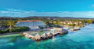 Aerial view of a coastal resort featuring multiple buildings, a wooden pier, and thatched-roof bungalows over clear blue waters, surrounded by lush greenery and rolling hills.