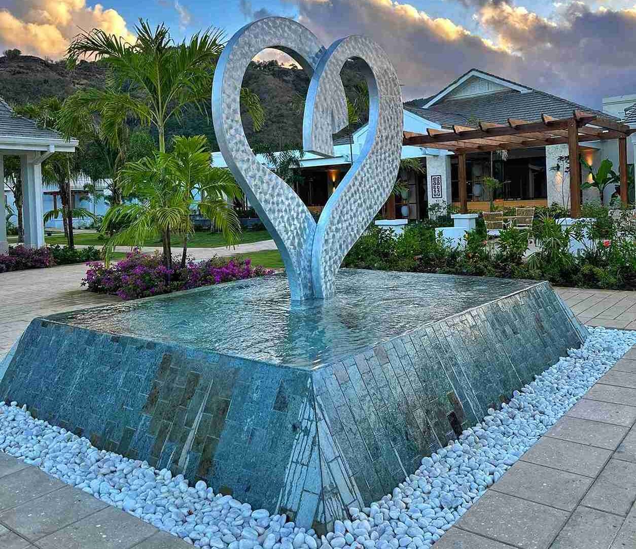 A large, metallic sculpture in the shape of a heart rises from a shallow water feature, surrounded by smooth white pebbles and lush tropical plants, set against a backdrop of a modern building and a cloudy sky.