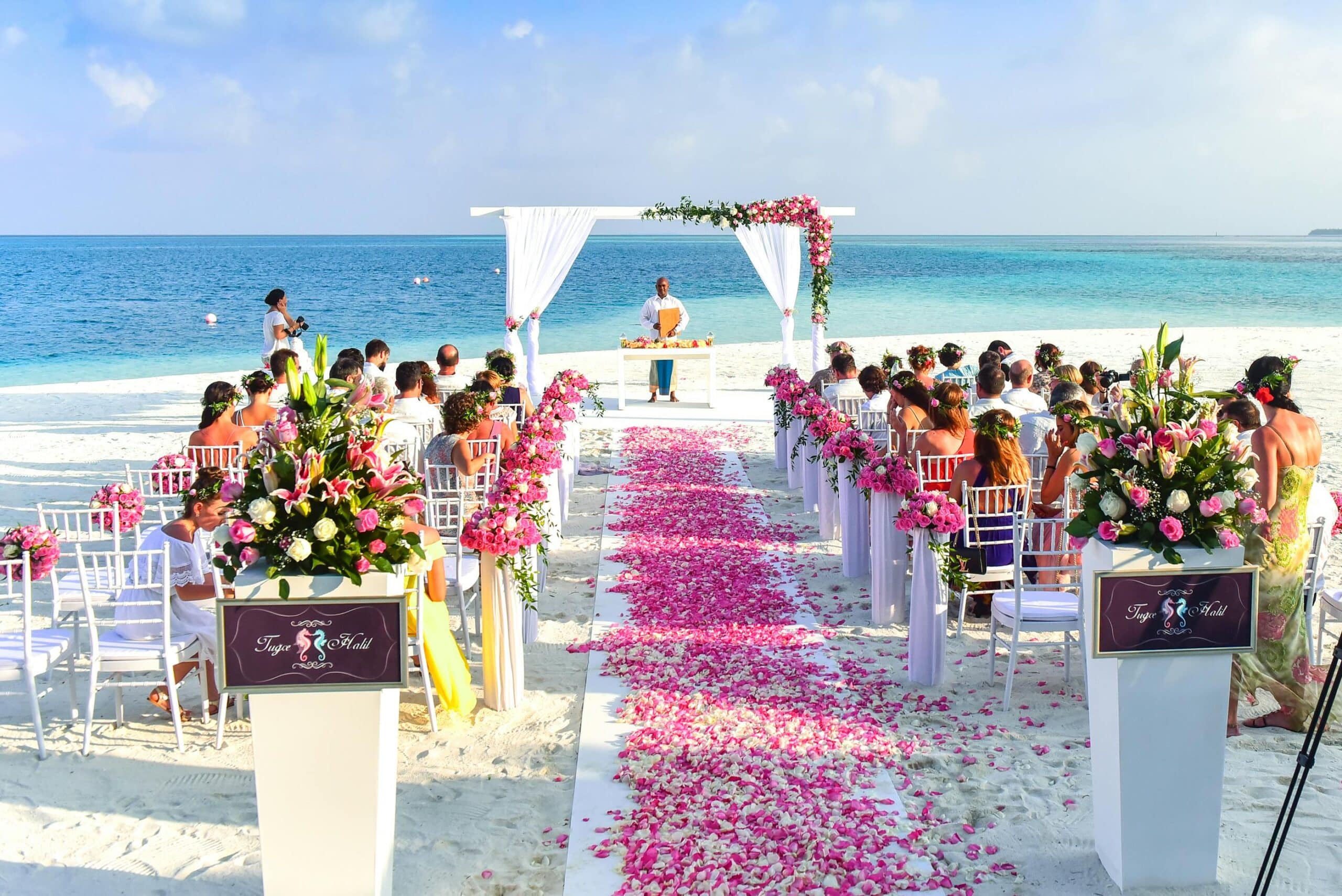 A picturesque beach wedding setup featuring a white aisle adorned with pink flower petals, flanked by elegantly arranged floral displays. Guests are seated on white chairs, with some wearing floral headbands, as an officiant stands under a floral arch by the shoreline, with a serene ocean backdrop and clear blue skies.