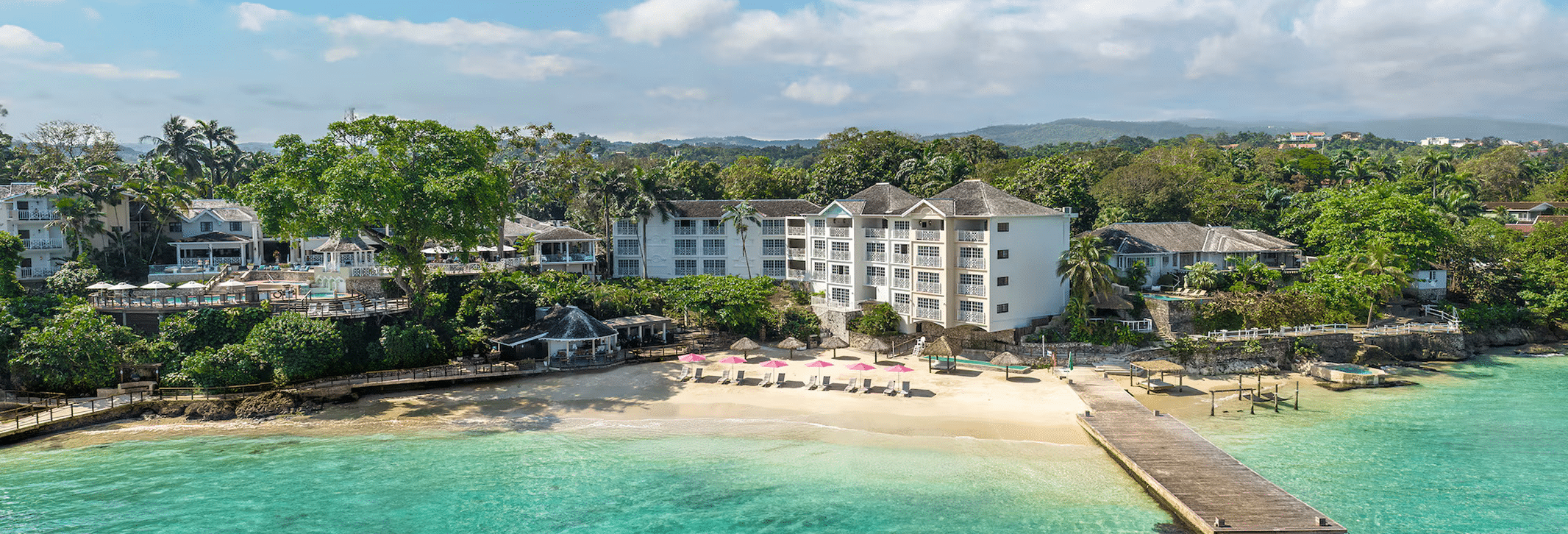 A scenic view of a coastal resort featuring white buildings, a beach with sandy shores, turquoise waters, and lush greenery. There are sun loungers with pink umbrellas on the beach and a wooden pier extending into the water.