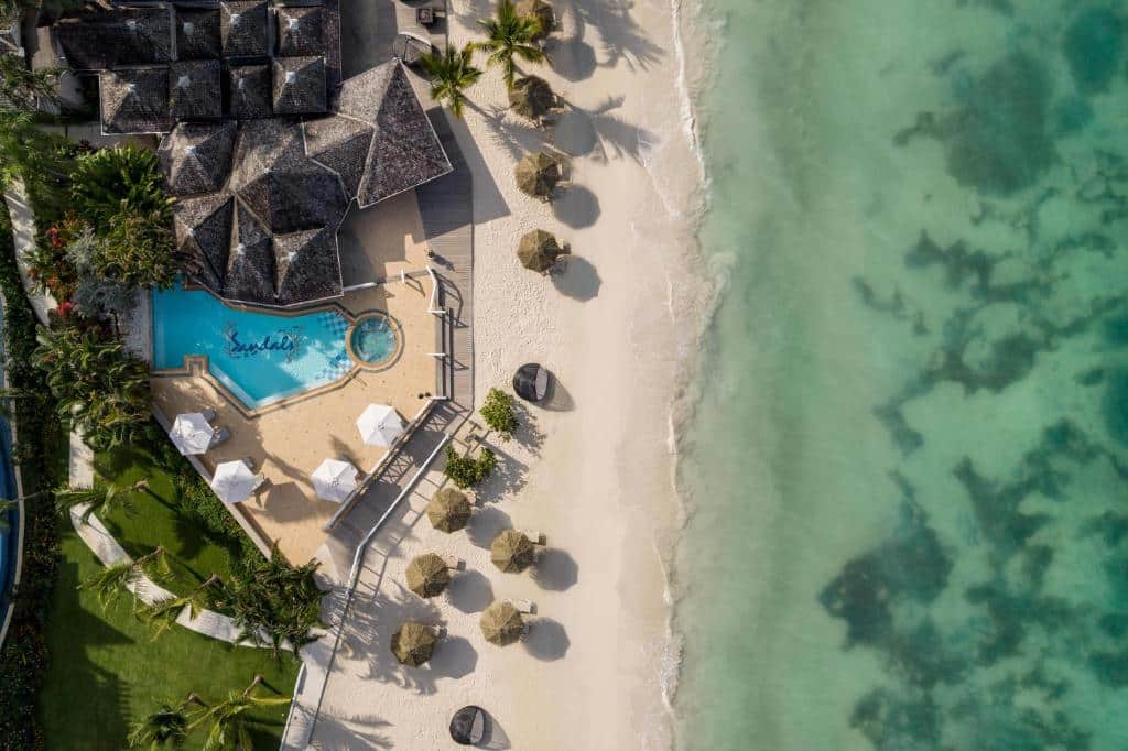 Aerial view of a beach resort featuring a swimming pool surrounded by lounge chairs and cabanas, with white sand beach and turquoise waters in the background.