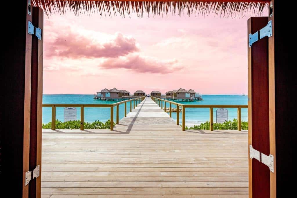 A view through open wooden doors framing a long wooden walkway leading to overwater bungalows on the horizon, with azure waters and a pastel pink sky dotted with clouds.