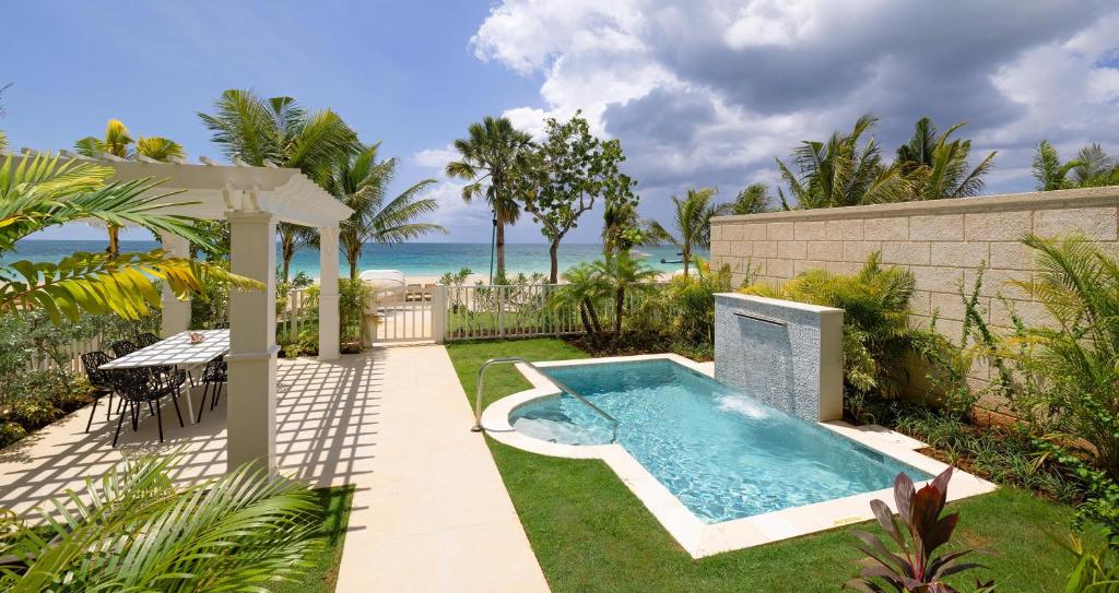 A tranquil outdoor setting featuring a small swimming pool with a water feature, surrounded by lush greenery and palm trees, with a patio area containing a dining table and chairs, overlooking a beach and ocean under a partly cloudy sky.
