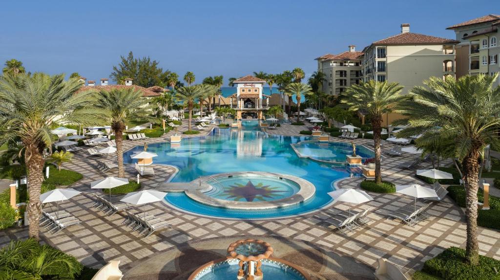 A luxurious resort pool area surrounded by palm trees, lounge chairs, and white umbrellas, with a decorative tiled pattern on the ground and buildings in the background under a clear blue sky.