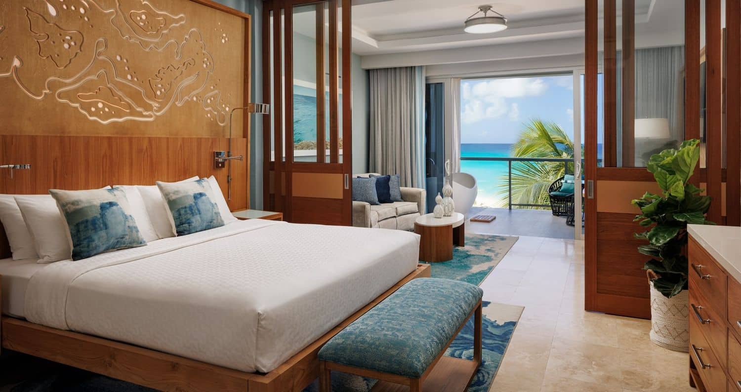 A modern hotel room featuring a king-sized bed with decorative pillows, a wooden headboard with intricate designs, and a cozy sitting area with a gray sofa. Large glass doors open to a balcony overlooking a turquoise ocean and palm trees, while a potted plant and a stylish dresser complete the room's elegant decor.