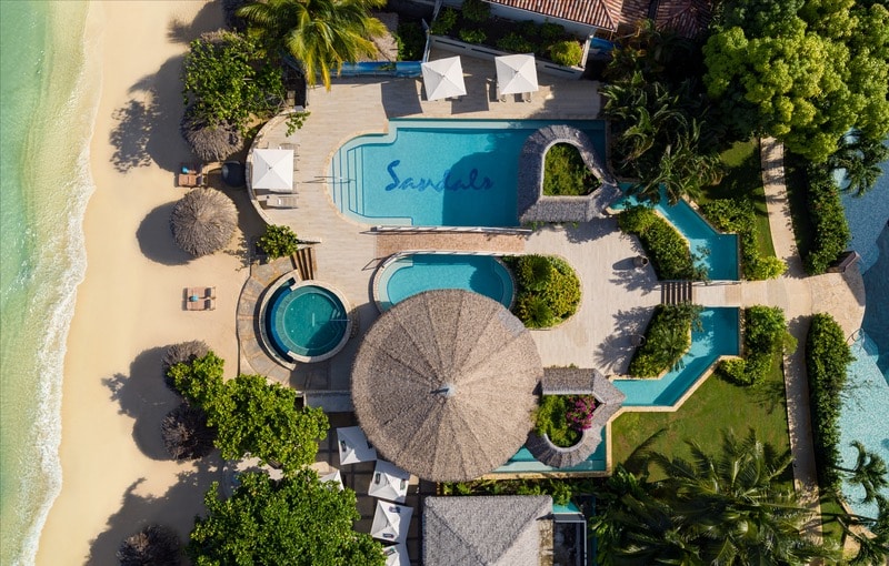 Aerial view of a luxurious resort featuring two pools, surrounded by lush greenery and beach loungers, with a sandy beach and ocean in the background. One pool has the word 'Sandals' elegantly displayed on the surface.