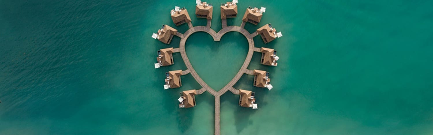 Aerial view of overwater bungalows arranged in a heart shape, surrounded by clear turquoise water.