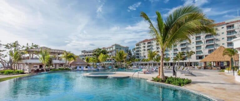 A luxurious resort pool area featuring a large swimming pool surrounded by palm trees, lounge chairs, and a thatched-roof pavilion, with a multi-story hotel building in the background and a clear blue sky above.