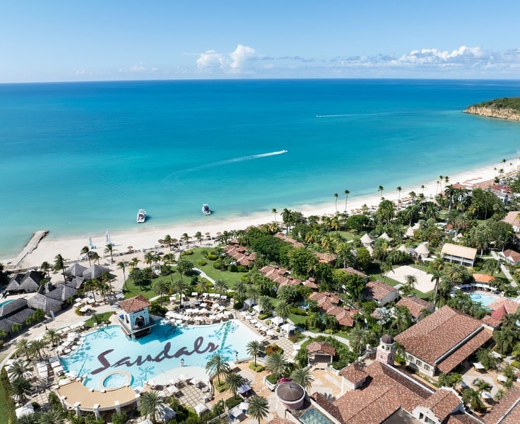 Aerial view of a tropical resort featuring a large, intricately designed pool with the word 'Sandals' written on the surface, lush green gardens, beach umbrellas, and white sand beach along a turquoise ocean.