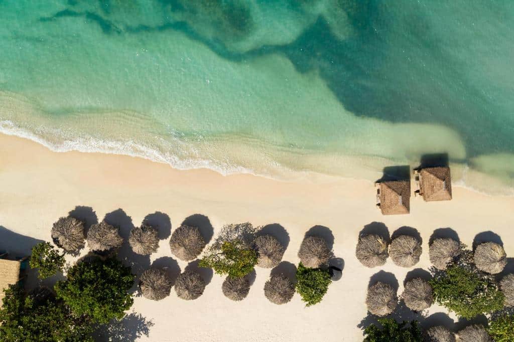 Aerial view of a serene beach with soft sand and clear turquoise water, featuring numerous thatched umbrellas lined up along the shore and two small wooden huts near the water's edge.