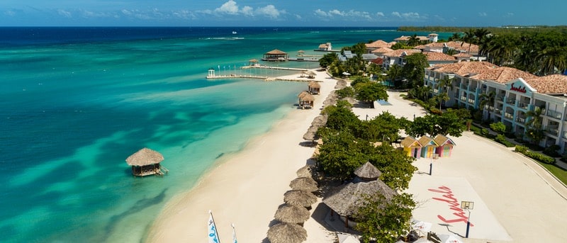 Aerial view of a tropical beach featuring turquoise waters, white sand, and palm trees. Beach huts and a wooden pier are visible in the background, along with a resort building and colorful beach houses near the shoreline.