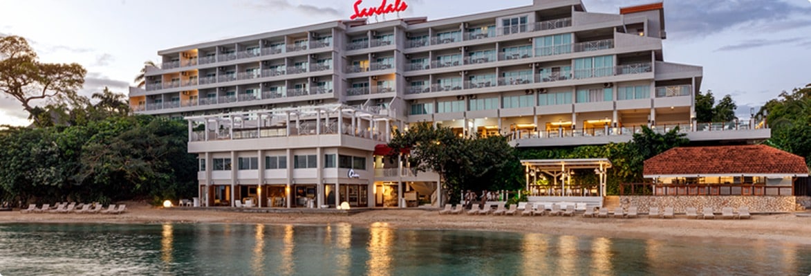 A coastal view of a Sandals resort hotel featuring a multi-story building with balconies, surrounded by lush greenery and a sandy beach. Lounge chairs are arranged along the shore, reflecting in the calm water at sunset.