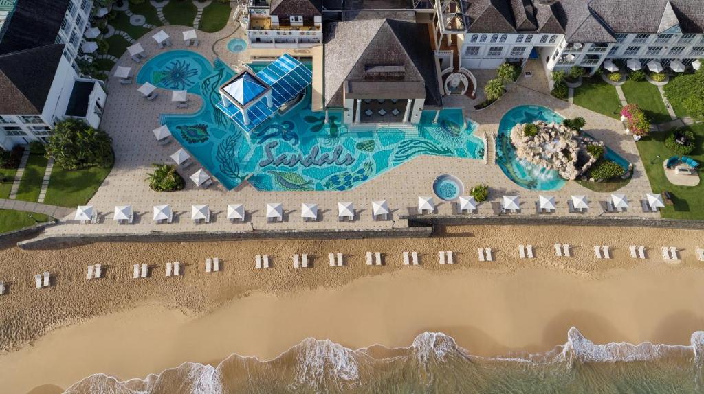 Aerial view of a luxurious beachfront resort with a large decorative swimming pool featuring artistic designs, surrounded by sun loungers and beach umbrellas, with soft sand and gentle waves in the foreground.