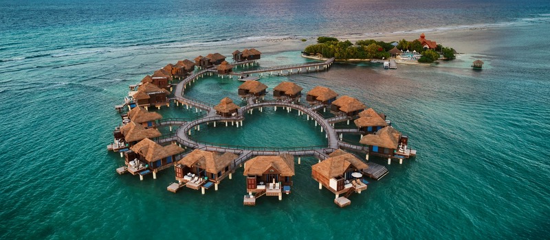 Aerial view of a heart-shaped arrangement of overwater bungalows surrounded by turquoise waters, with a small island and lush vegetation in the background.