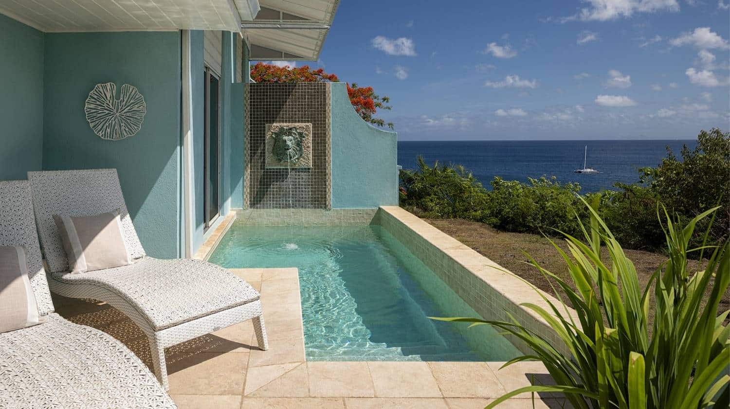 A luxurious patio with two white lounge chairs beside a small private infinity pool overlooking the ocean, featuring a decorative wall with a water fountain, set against a backdrop of blue skies and scattered clouds.