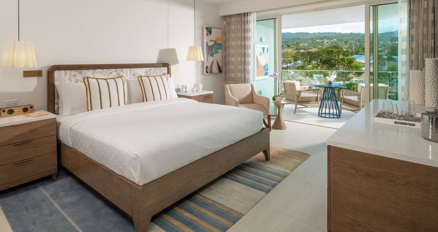 A stylish hotel room featuring a queen-sized bed with decorative pillows, a wooden nightstand, and a modern armchair. The room has large windows overlooking a scenic outdoor area with greenery, and there's a small balcony with outdoor seating. The decor includes a colorful painting and soft lighting, creating a cozy atmosphere.