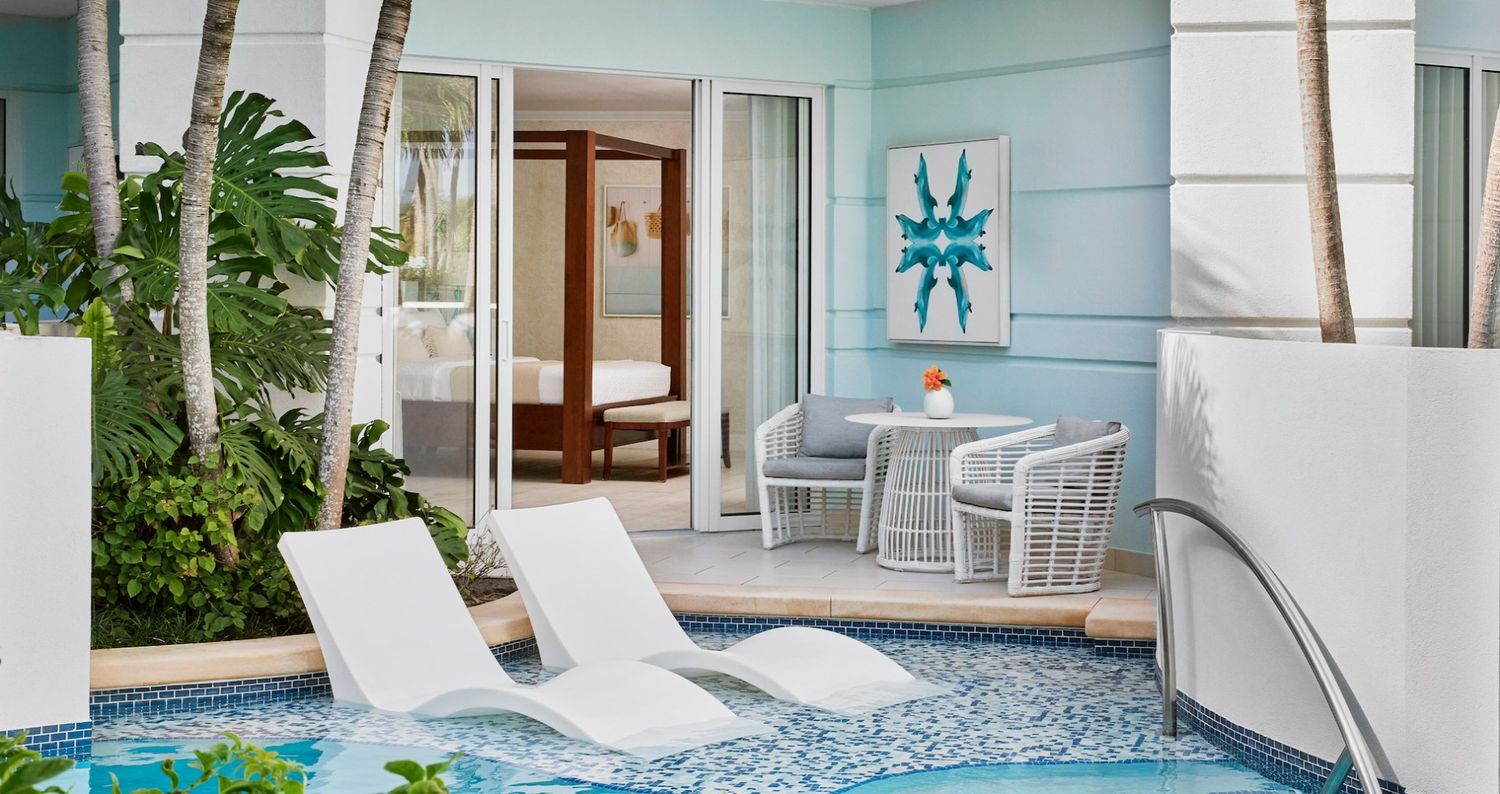 A serene outdoor area featuring a small pool with white lounge chairs, surrounded by greenery and palm trees. In the background, a glass door leads to a room with a wooden four-poster bed and a small round table with two wicker chairs. The walls are painted a light blue.