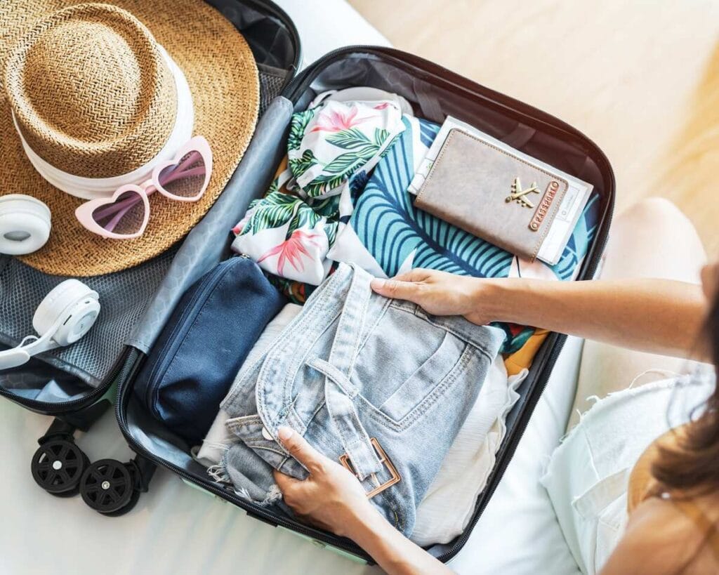 An open suitcase filled with neatly packed items, including a straw hat, pink heart-shaped sunglasses, a pair of white headphones, colorful clothing, a dark blue pouch, and a beige travel wallet. A hand is seen adjusting a pair of light denim shorts in the suitcase.