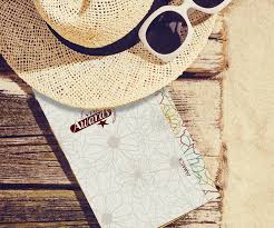 A straw sun hat rests on a wooden surface next to a pair of white sunglasses and a coloring book, which has a colorful floral design on its cover and is partially open, suggesting a relaxing outdoor atmosphere.