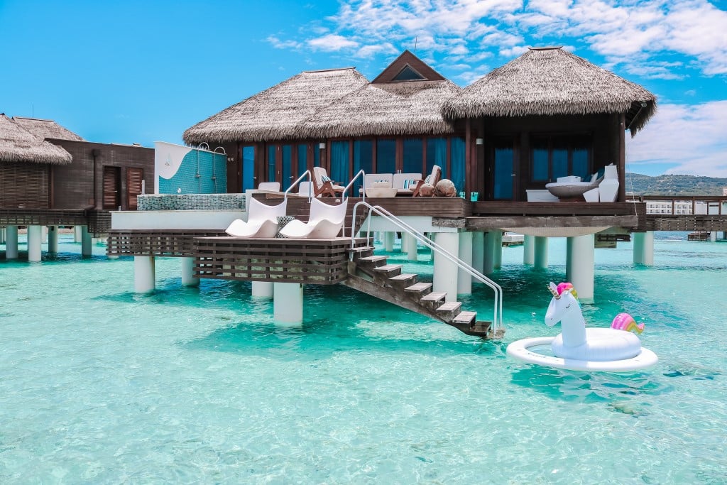 A picturesque overwater bungalow with a thatched roof, surrounded by clear turquoise water, featuring a small staircase and two white lounge chairs on the deck. A unicorn-shaped inflatable float is visible in the water nearby.