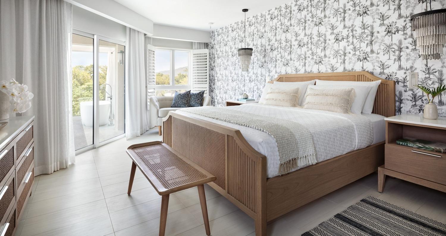 A stylish bedroom featuring a wooden bed with white linens and decorative pillows, a comfortable lounge chair with patterned cushions, and light-colored walls with floral wallpaper. A glass door leads to a small balcony, and a decorative light fixture hangs from the ceiling. The room includes a wooden bench at the foot of the bed and a nightstand with a plant and a book.